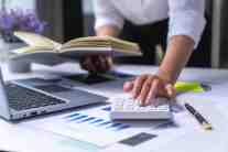 Close-up of a professional's hand using a white calculator at a desk with an open laptop, financial chart documents, a pen, and a book.