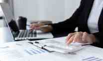 Professional reviewing financial reports and charts while using a calculator and laptop at a desk.