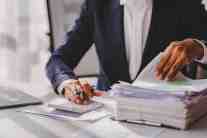 A person in a suit using a calculator and reviewing stacks of financial papers and charts.