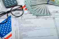 An IRS Form 1120 corporate tax return document laid out on a desk with a fanned stack of 100-dollar bills, eyeglasses, a calculator, and a US flag banner.
