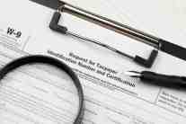 An official IRS Form W-9 attached to a black clipboard with a fountain pen and magnifying glass resting on top.