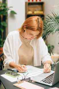 Red-haired woman filling out a form at a desk with a laptop, calculator, glasses, and a drink nearby.