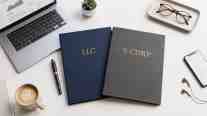 Two portfolios embossed with "LLC" and "S-Corp" laying on a white desk  with a laptop, a cell phone, a cup of coffee, and eye glasses.